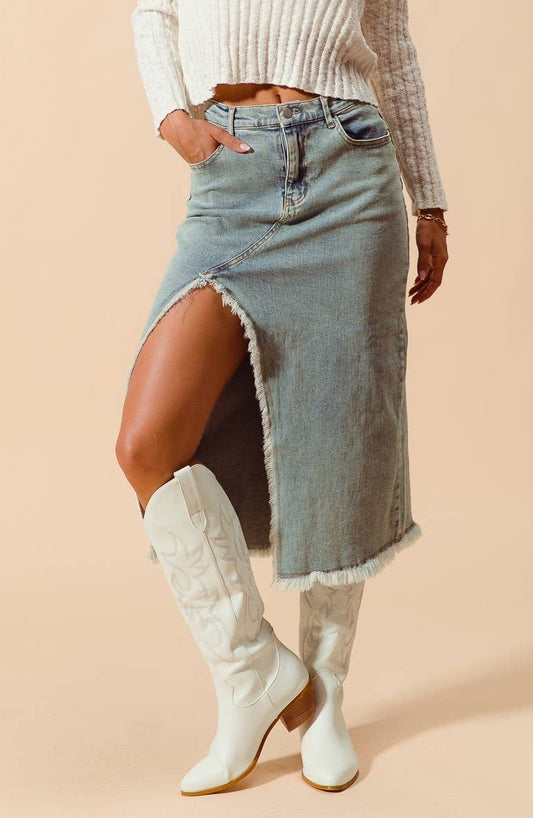 A model wearing a light-wash denim midi skirt with a high side slit and frayed hem, paired with white western-style boots and a cream ribbed sweater, set against a solid beige background.