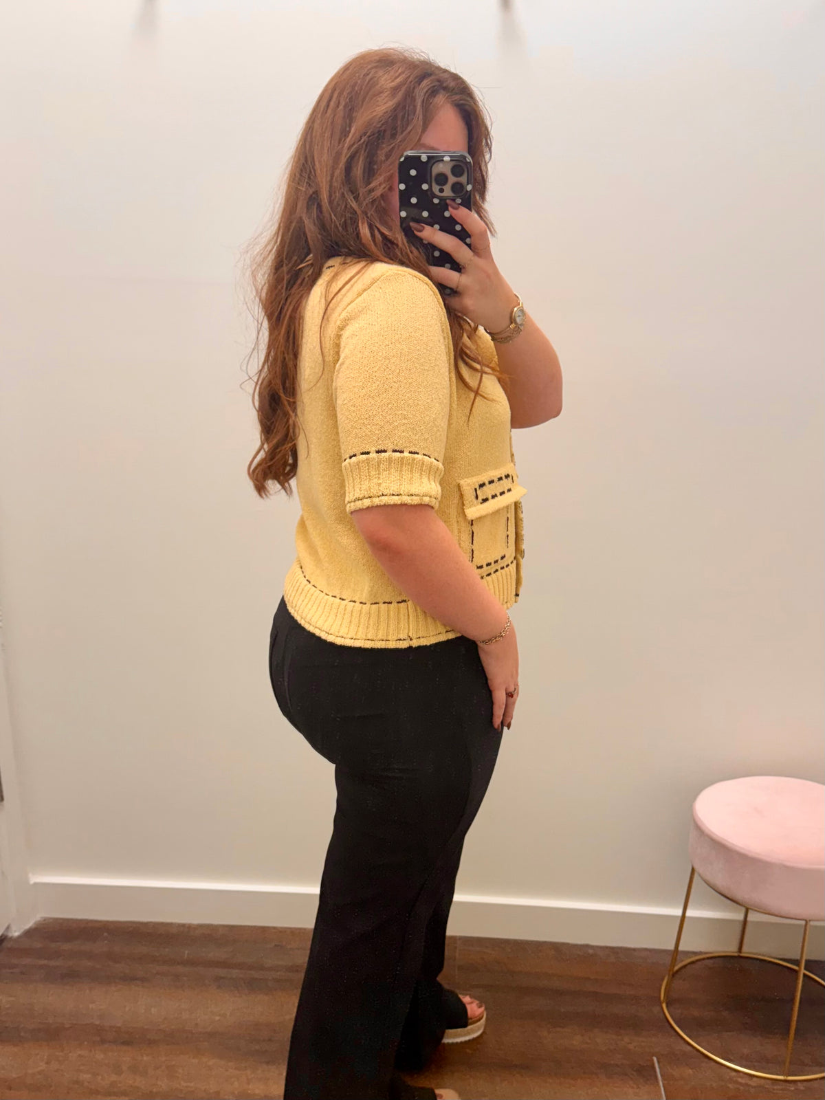 A woman takes a mirror selfie in a fitting room wearing a pale yellow knit short-sleeve top with decorative black trim, paired with black pants.
