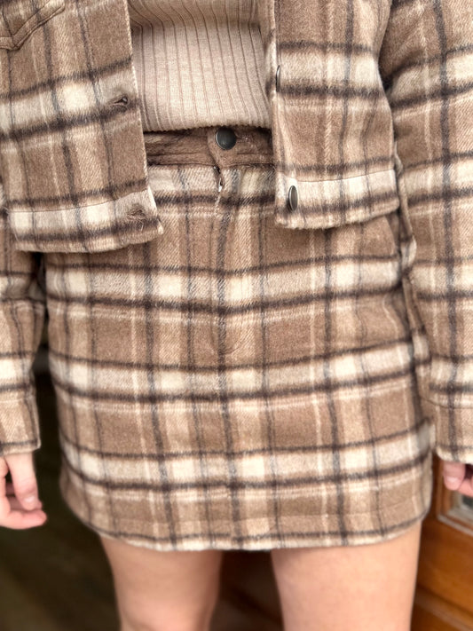 A model showing off a close up view of a brown, khaki, & cream plaid skirt.
