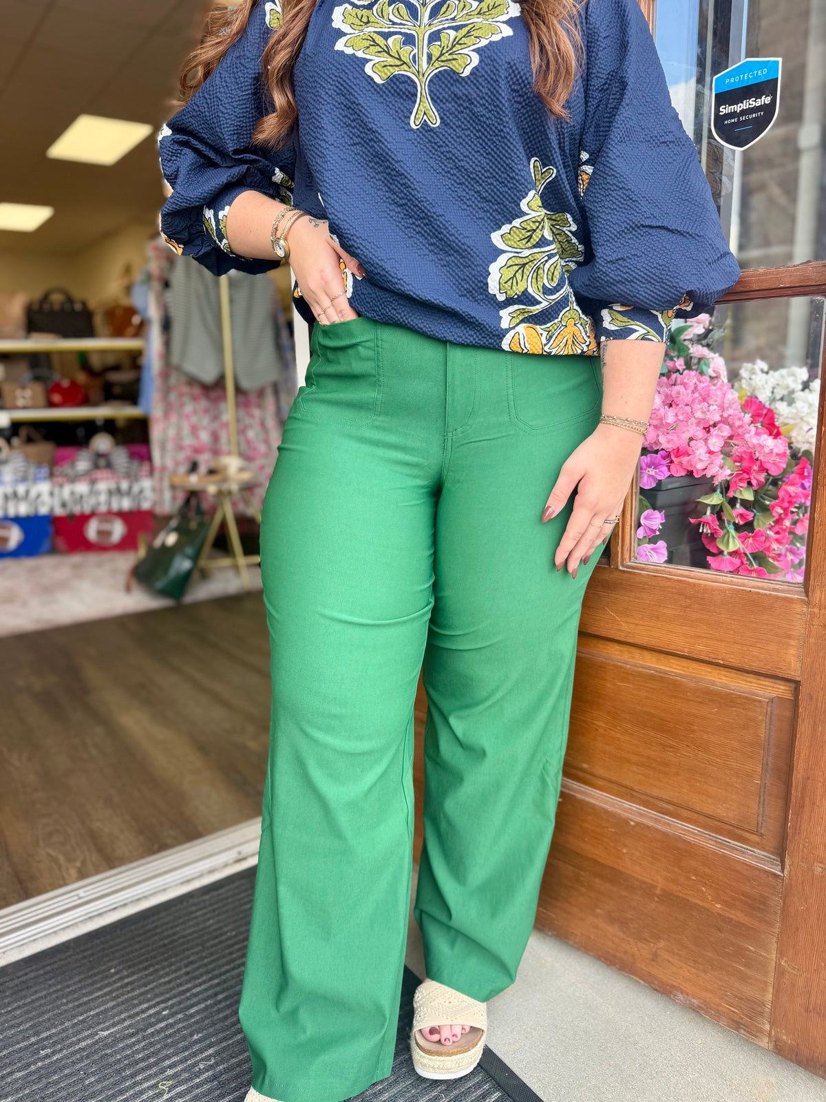A person standing in a storefront wearing bright green wide-leg pants and a navy blue textured blouse with a yellow and green floral pattern. The photo is cropped from the shoulders down, showing the pants and lower torso. 