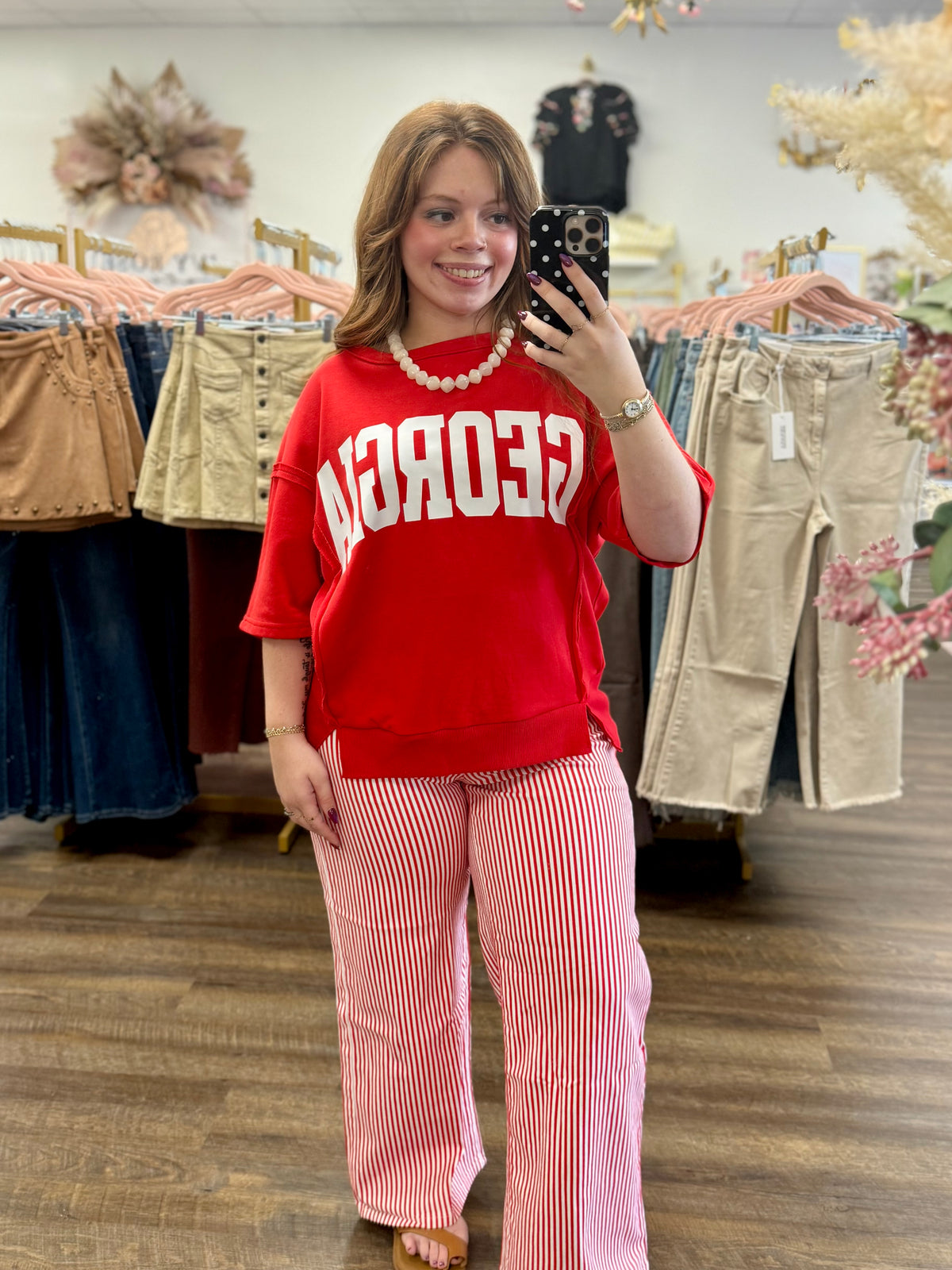 A woman takes a mirror selfie in a clothing boutique wearing a red sweatshirt with white 'GEORGIA' lettering and red-and-white striped wide-leg pants.
