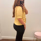 A woman takes a mirror selfie in a fitting room wearing a pale yellow knit short-sleeve top with decorative black trim, paired with black pants.