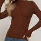 A woman wearing a brown ribbed mock neck long-sleeve sweater and blue jeans, posing against a neutral beige background.