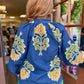 A woman viewed from behind wearing a blue textured blouse with large yellow and green floral patterns, paired with bright green pants, standing in front of a shop window.