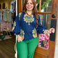 A woman stands in the doorway of a boutique wearing a navy blue long-sleeve blouse with a yellow and green floral pattern and bright green pants.