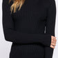 A woman wearing a black ribbed turtleneck sweater and blue jeans, standing against a plain light gray studio background.