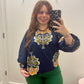 A woman taking a mirror selfie wearing a navy blue long-sleeve blouse with yellow and green floral designs, paired with green pants.