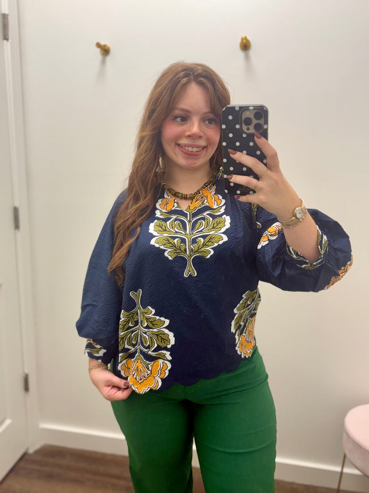 A woman taking a mirror selfie wearing a navy blue long-sleeve blouse with yellow and green floral designs, paired with green pants.