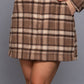 A woman wearing a brown and beige plaid mini skirt with a matching long-sleeve top, against a solid gray studio background.