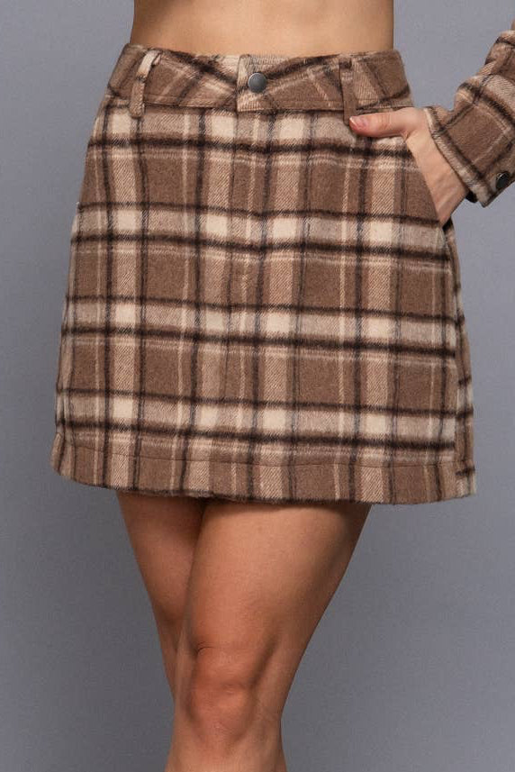 A woman wearing a brown and beige plaid mini skirt with a matching long-sleeve top, against a solid gray studio background.