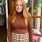 A woman stands in front of a wooden window, wearing a brown ribbed turtleneck sweater and a brown and beige plaid skirt.