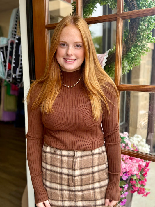 A woman stands in front of a wooden window, wearing a brown ribbed turtleneck sweater and a brown and beige plaid skirt.