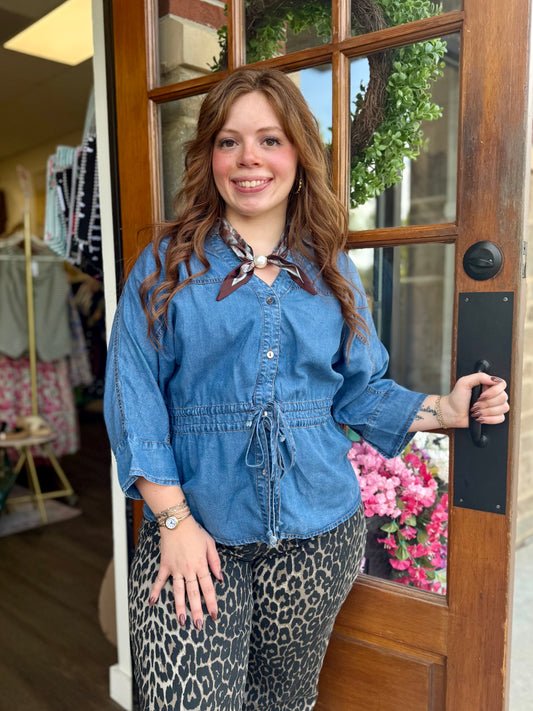 A woman stands in a doorway wearing a blue denim long-sleeve top with a drawstring waist and a patterned scarf, paired with leopard-print pants.