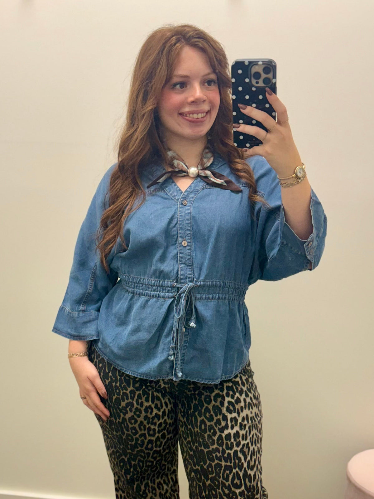 A woman taking a mirror selfie wearing a blue denim button-up shirt with a drawstring waist and a patterned neck scarf, paired with leopard print pants.