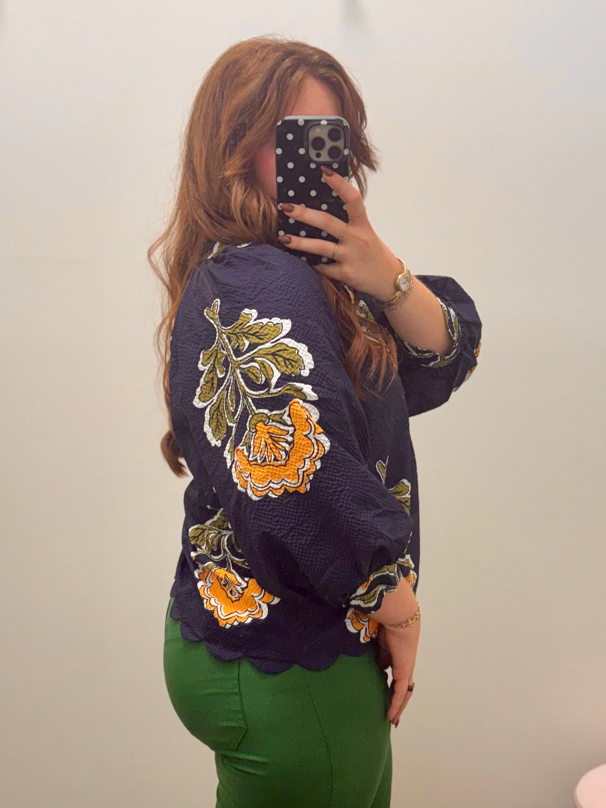 A woman taking a mirror selfie wearing a navy blue textured blouse with yellow and green floral embroidery on the sleeves and bodice, paired with green pants.
