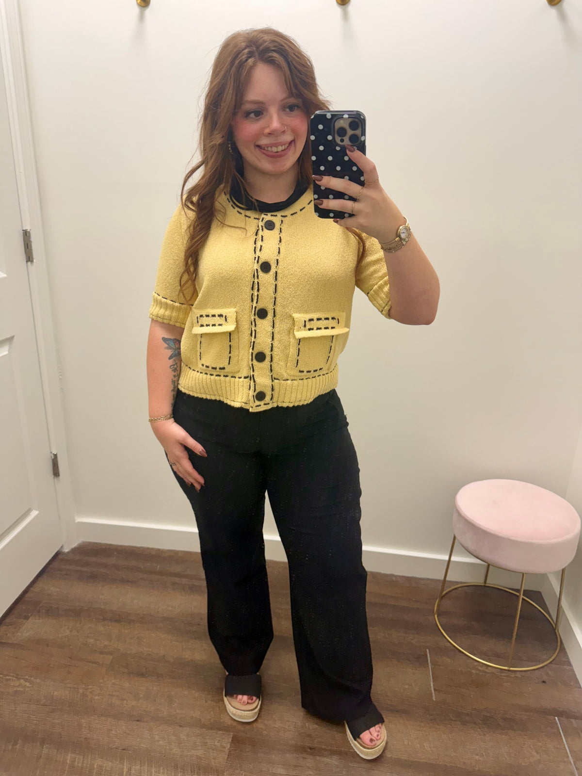 A woman takes a mirror selfie wearing a yellow short-sleeve knit cardigan with black buttons and trim, paired with black wide-leg pants. 