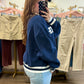 Mirror selfie in a clothing boutique showing the side view of a navy blue varsity-style sweatshirt with white stripes.