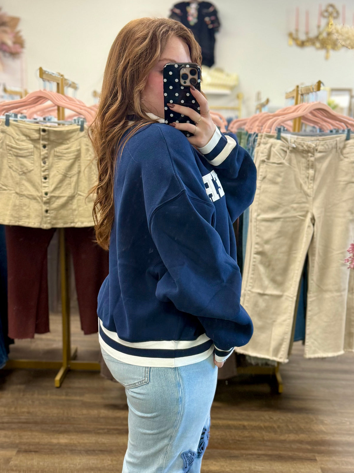 Mirror selfie in a clothing boutique showing the side view of a navy blue varsity-style sweatshirt with white stripes.