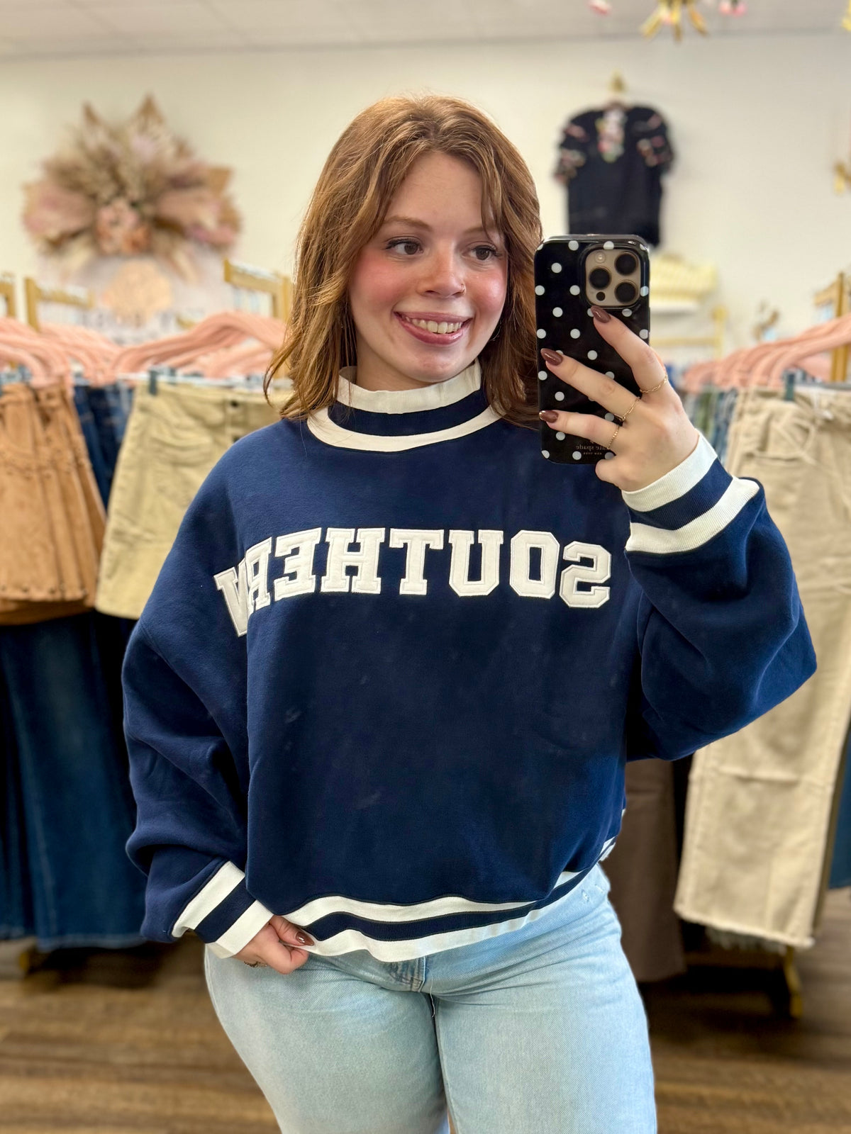 Mirror selfie taken in a clothing boutique, wearing a navy blue varsity-style sweatshirt with 'SOUTHERN' printed in white across the chest and light blue jeans.