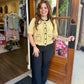 A woman stands in a storefront doorway wearing a pale yellow short-sleeve knit top with black trim and buttons, paired with black high-rise pants. 
