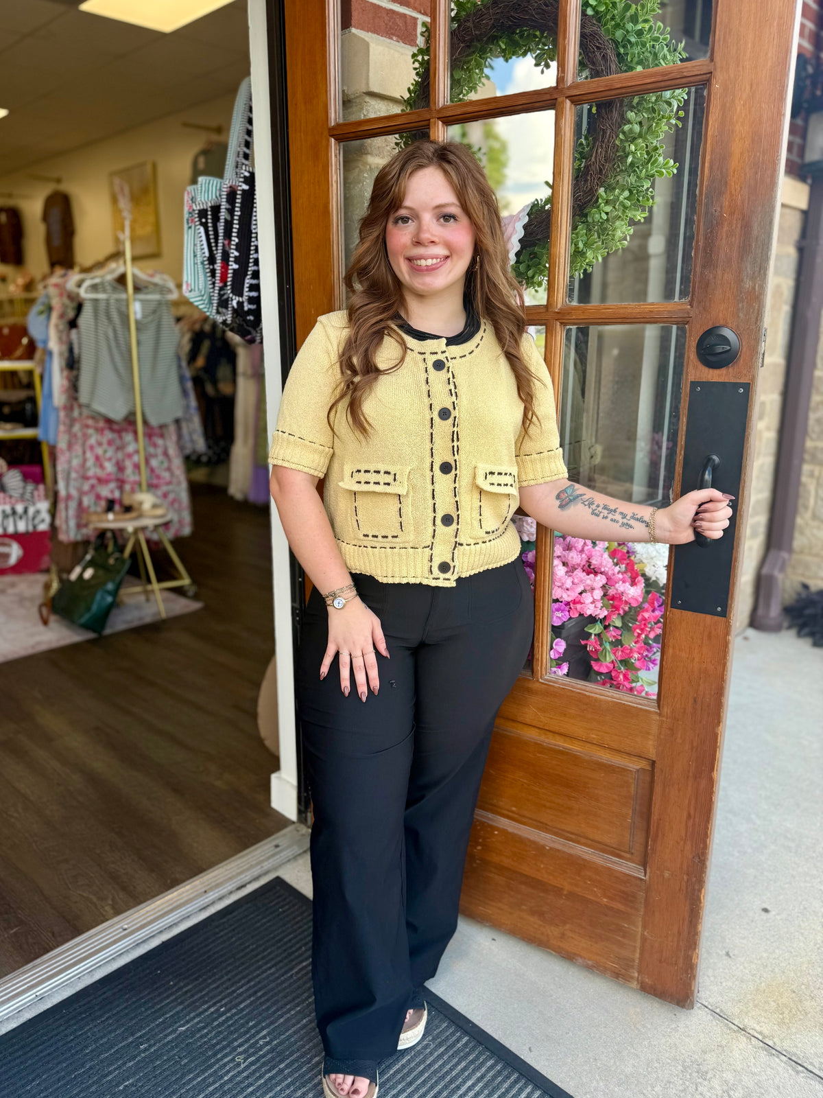 A woman stands in a storefront doorway wearing a pale yellow short-sleeve knit top with black trim and buttons, paired with black high-rise pants. 