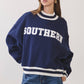 A woman wearing a navy blue crewneck sweatshirt with 'SOUTHERN' printed in white varsity letters across the chest, paired with blue jeans, against a plain light gray studio background.