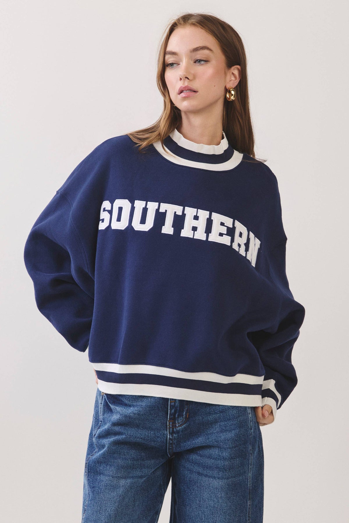 A woman wearing a navy blue crewneck sweatshirt with 'SOUTHERN' printed in white varsity letters across the chest, paired with blue jeans, against a plain light gray studio background.