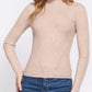 A woman wearing a beige ribbed mock neck long-sleeve sweater and blue jeans, standing against a plain white studio background.