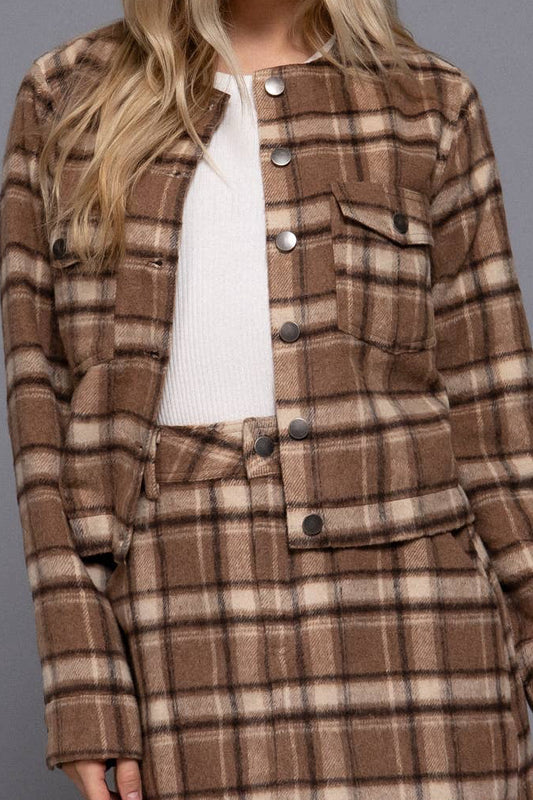 A model wearing a brown and beige plaid button-up jacket and matching mini skirt over a white top, photographed against a solid gray studio background.